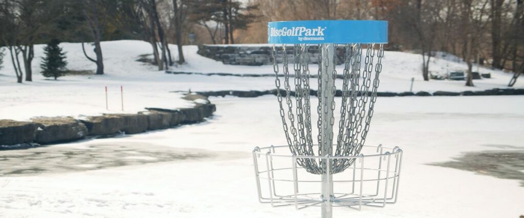 A disc golf basket set in the foreground of a winter scene on a golf course with a water feature.