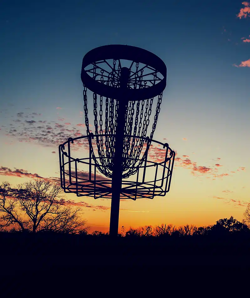 A disc golf basket silhouetted by an orange and yellow sunset sky.